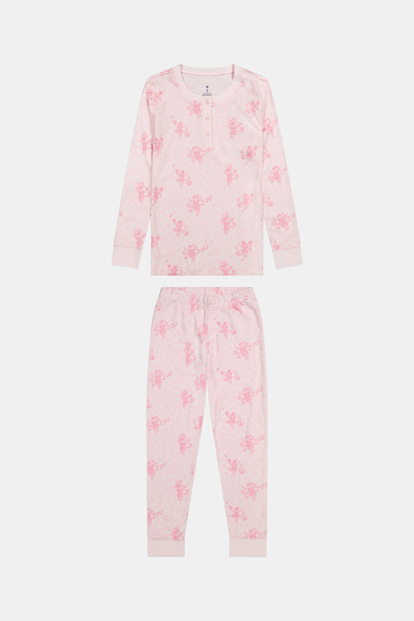 A girl with long brown hair stands against a pale pink wall, wearing Roller Rabbit Kids Cupid's Kiss Juniper Pajamas in light pink and gray socks, looking at the camera with a slight smile.