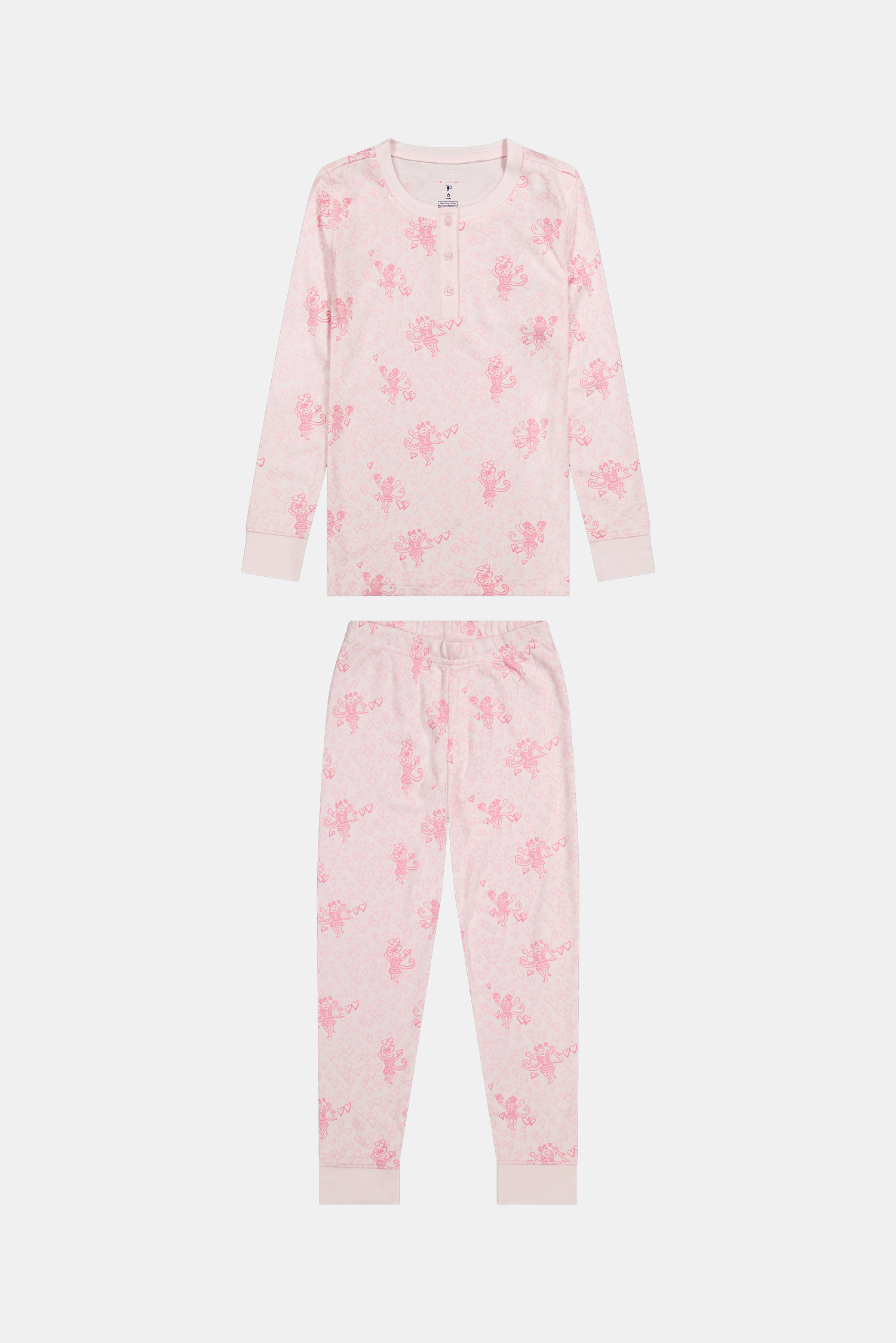 A girl with long brown hair stands against a pale pink wall, wearing Roller Rabbit Kids Cupid's Kiss Juniper Pajamas in light pink and gray socks, looking at the camera with a slight smile.