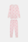 A girl with long brown hair stands against a pale pink wall, wearing Roller Rabbit Kids Cupid's Kiss Juniper Pajamas in light pink and gray socks, looking at the camera with a slight smile.
