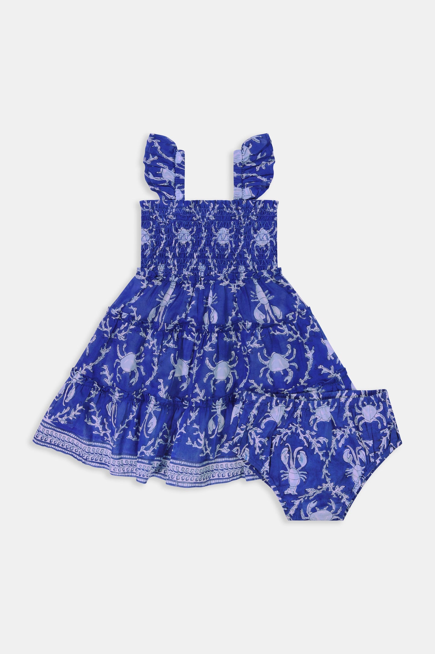 A young girl with straight brown hair smiles while wearing the Roller Rabbit Kids Lobster Trellis Ceri Dress, featuring a blue and white pattern with ruffled sleeves, against a plain white background.