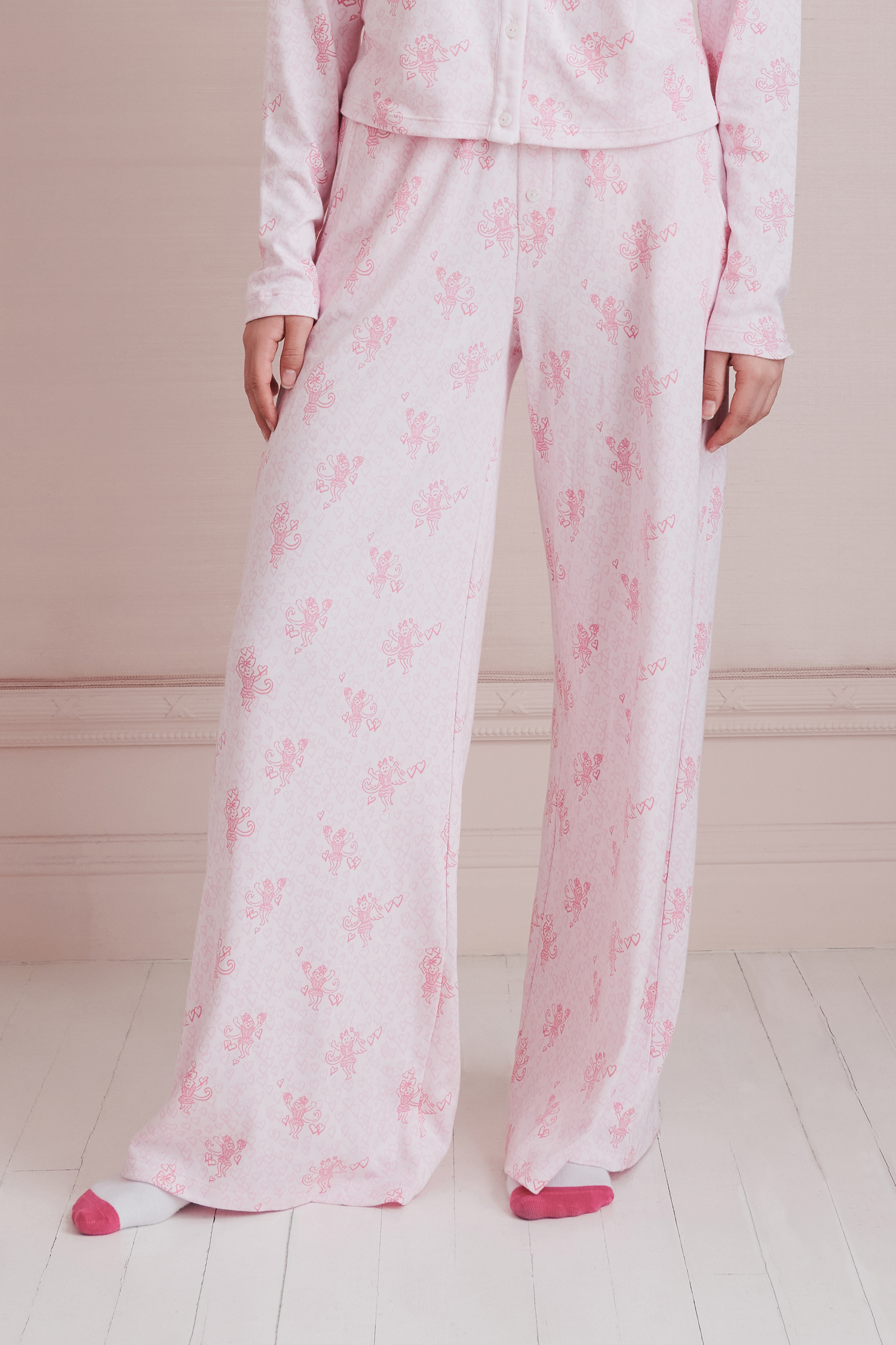 A young woman wearing the Roller Rabbit Cupid's Kiss Boden Pant and fluffy slippers stands in front of a pastel pink, candy-themed counter adorned with jars of sweets and heart decorations.