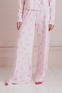A young woman wearing the Roller Rabbit Cupid's Kiss Boden Pant and fluffy slippers stands in front of a pastel pink, candy-themed counter adorned with jars of sweets and heart decorations.