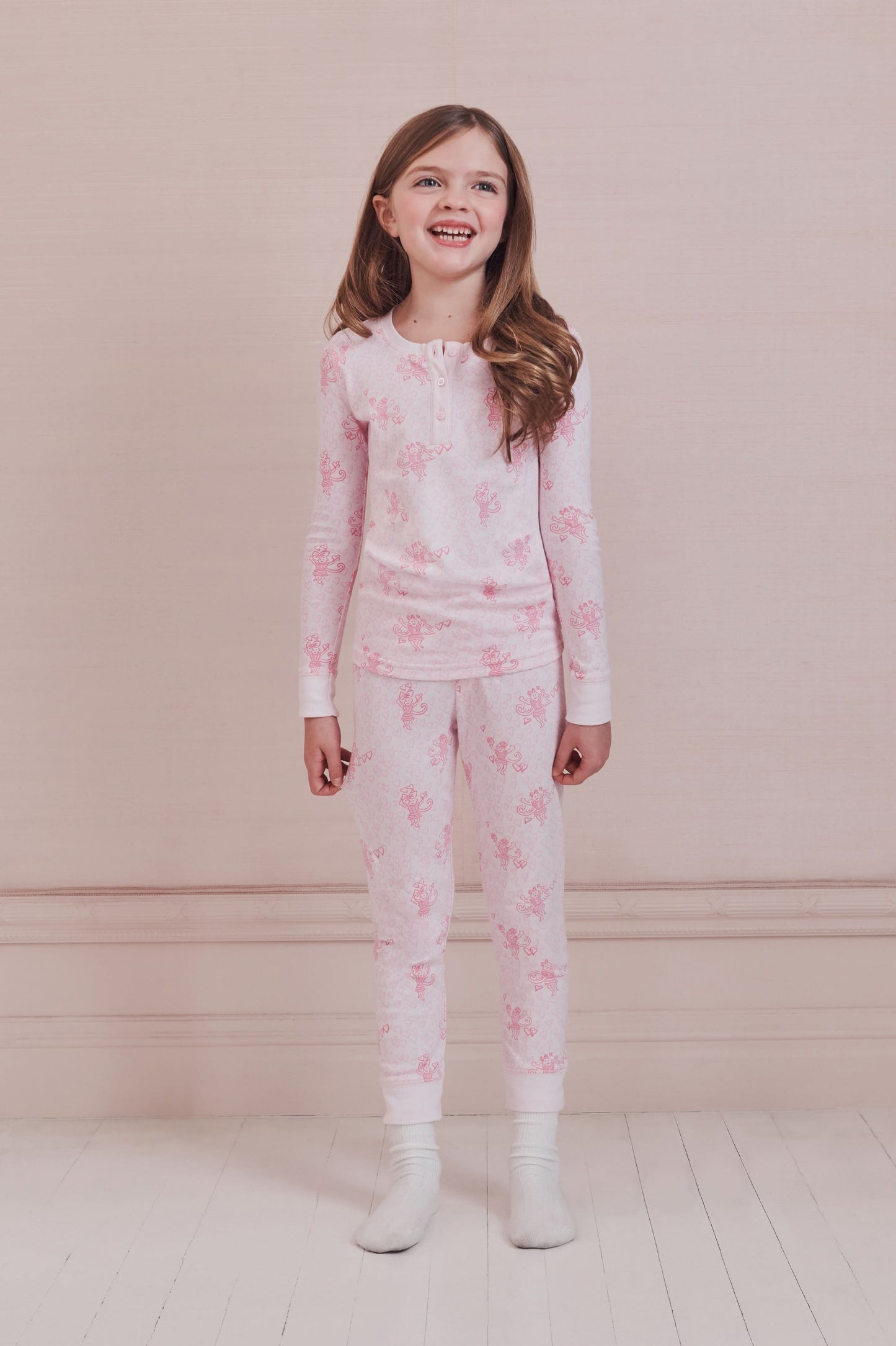 A girl with long brown hair stands against a pale pink wall, wearing Roller Rabbit Kids Cupid's Kiss Juniper Pajamas in light pink and gray socks, looking at the camera with a slight smile.