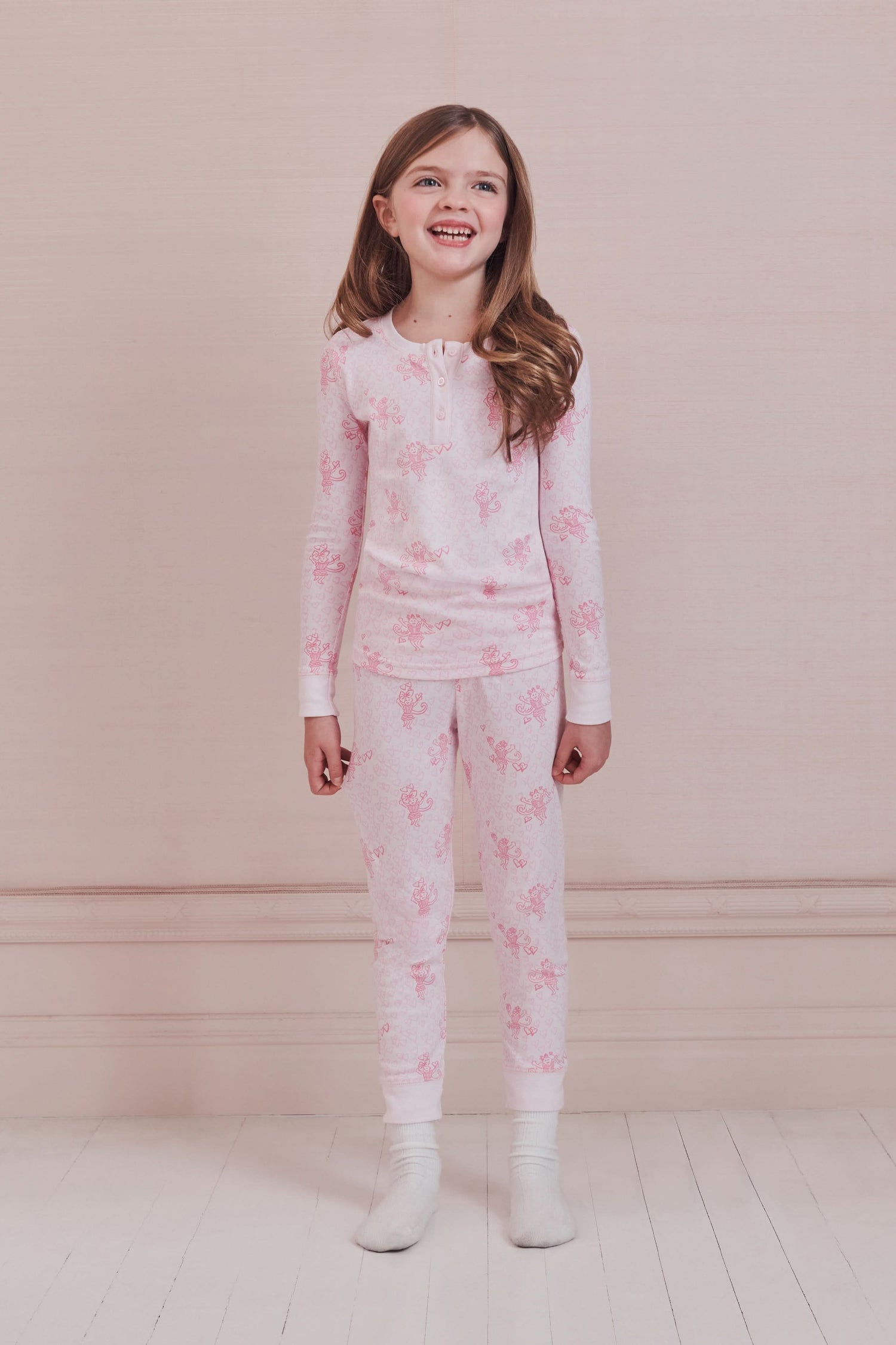 A girl with long brown hair stands against a pale pink wall, wearing Roller Rabbit Kids Cupid's Kiss Juniper Pajamas in light pink and gray socks, looking at the camera with a slight smile.