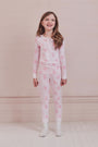 A girl with long brown hair stands against a pale pink wall, wearing Roller Rabbit Kids Cupid's Kiss Juniper Pajamas in light pink and gray socks, looking at the camera with a slight smile.