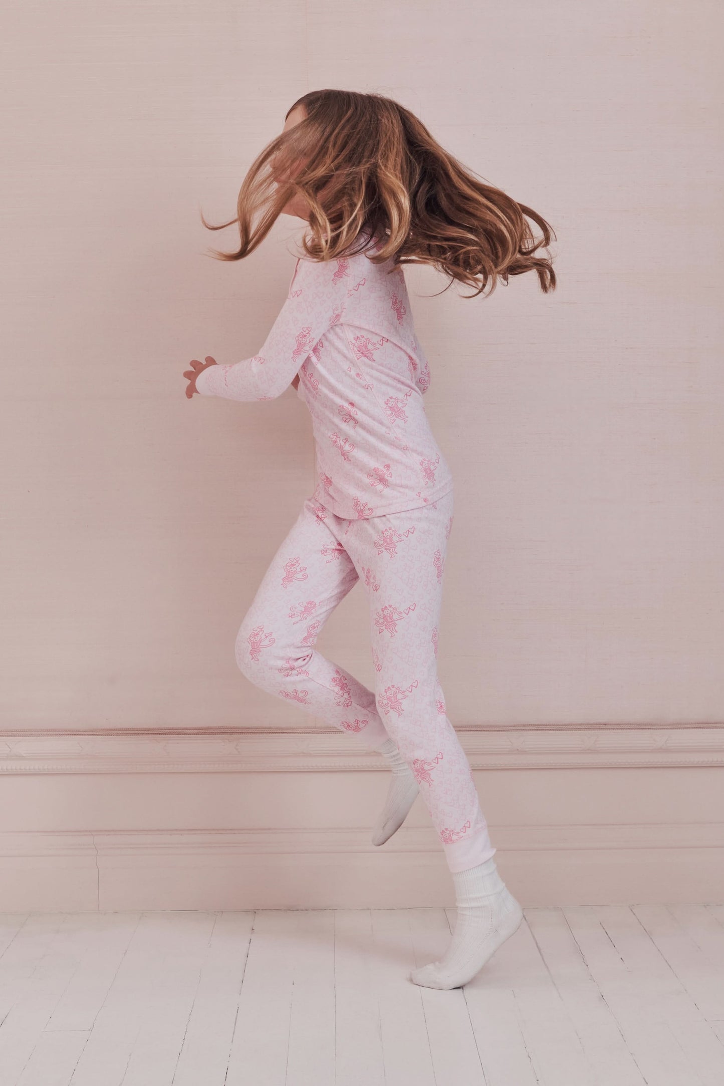 A girl with long brown hair stands against a pale pink wall, wearing Roller Rabbit Kids Cupid's Kiss Juniper Pajamas in light pink and gray socks, looking at the camera with a slight smile.