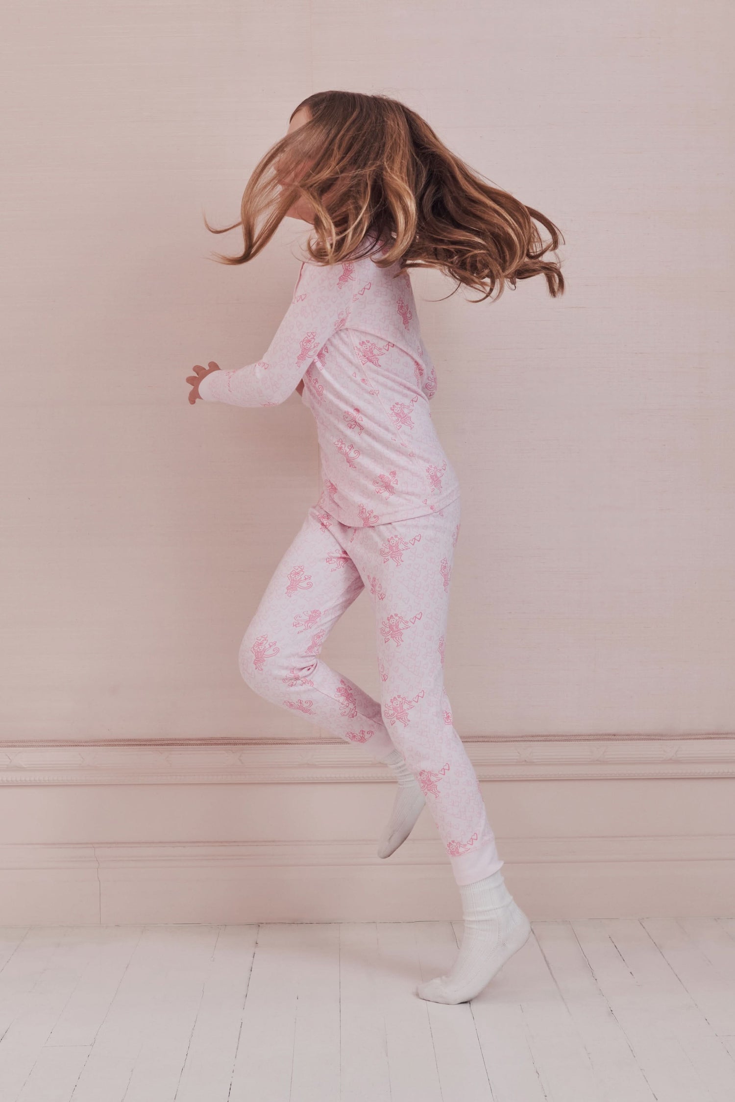 A girl with long brown hair stands against a pale pink wall, wearing Roller Rabbit Kids Cupid's Kiss Juniper Pajamas in light pink and gray socks, looking at the camera with a slight smile.