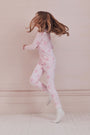 A girl with long brown hair stands against a pale pink wall, wearing Roller Rabbit Kids Cupid's Kiss Juniper Pajamas in light pink and gray socks, looking at the camera with a slight smile.