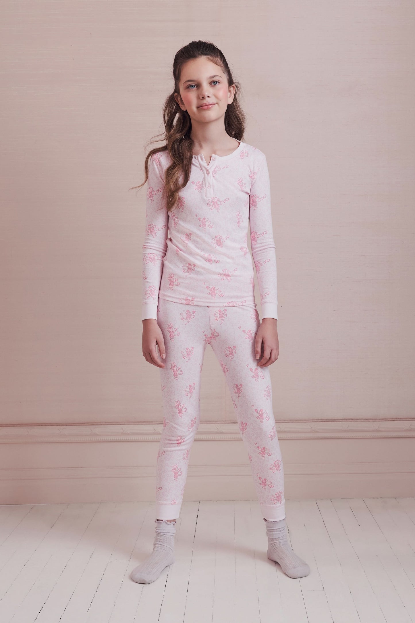 A girl with long brown hair stands against a pale pink wall, wearing Roller Rabbit Kids Cupid's Kiss Juniper Pajamas in light pink and gray socks, looking at the camera with a slight smile.