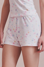 Wearing Roller Rabbit's Candy Heart Stripe Jones Shortie pajama shorts, featuring a pastel hearts, bows, and stars pattern, this person stands against a light background.