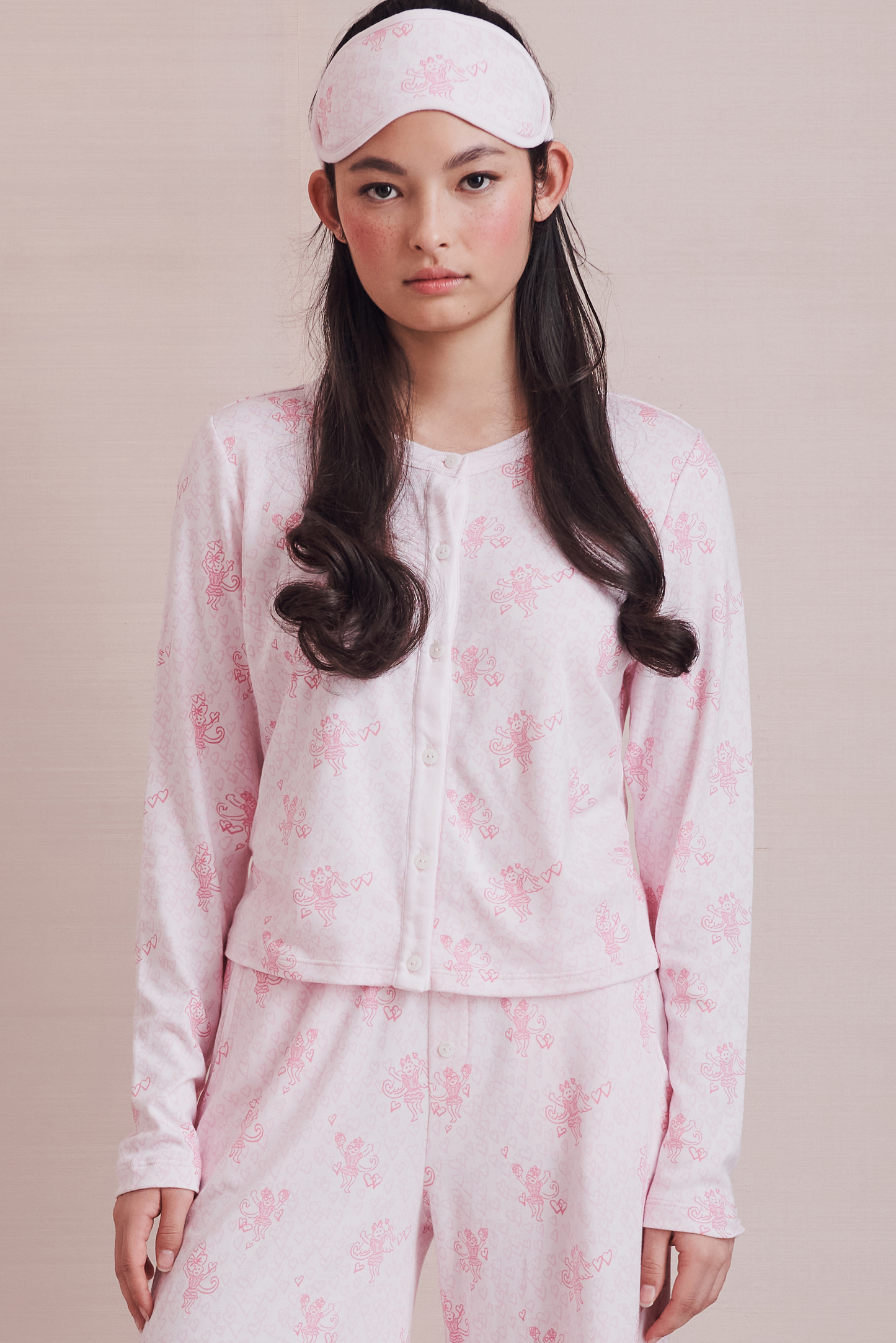 Wearing the Cupid's Kiss Nessa Cardigan by Roller Rabbit, a woman stands against a plain light background. The soft cardigan features a charming pink and white design with a delicate floral pattern, perfectly complemented by her matching sleep mask.