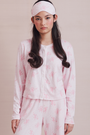 Wearing the Cupid's Kiss Nessa Cardigan by Roller Rabbit, a woman stands against a plain light background. The soft cardigan features a charming pink and white design with a delicate floral pattern, perfectly complemented by her matching sleep mask.