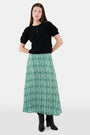 Someone wears the Roller Rabbit Fern Floral Megan Skirt—a green and white pleated maxi—paired with a black top, gold bracelets, and black heeled boots, shown from the waist down against a plain background.