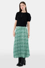 Someone wears the Roller Rabbit Fern Floral Megan Skirt—a green and white pleated maxi—paired with a black top, gold bracelets, and black heeled boots, shown from the waist down against a plain background.