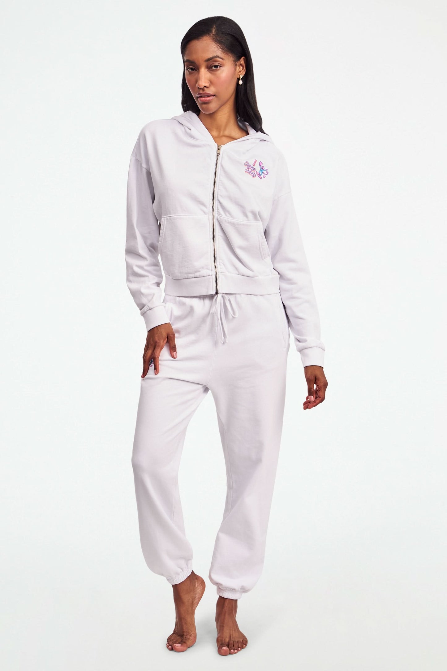 A person stands barefoot in Roller Rabbit O.C. Monkey Sweatpants and a matching hoodie, featuring colorful embroidered and printed graphics near the left pocket. The background is plain white.