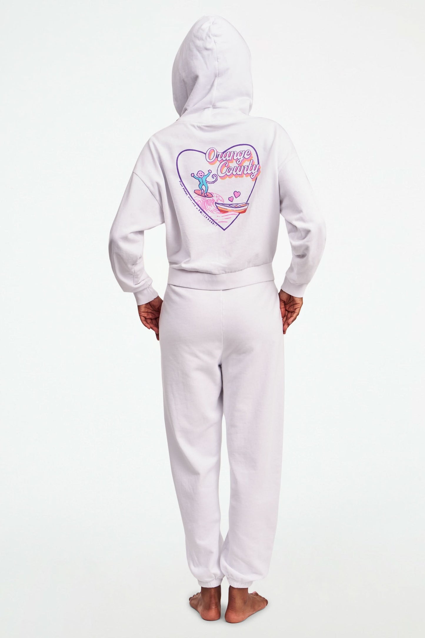 A person stands barefoot in Roller Rabbit O.C. Monkey Sweatpants and a matching hoodie, featuring colorful embroidered and printed graphics near the left pocket. The background is plain white.