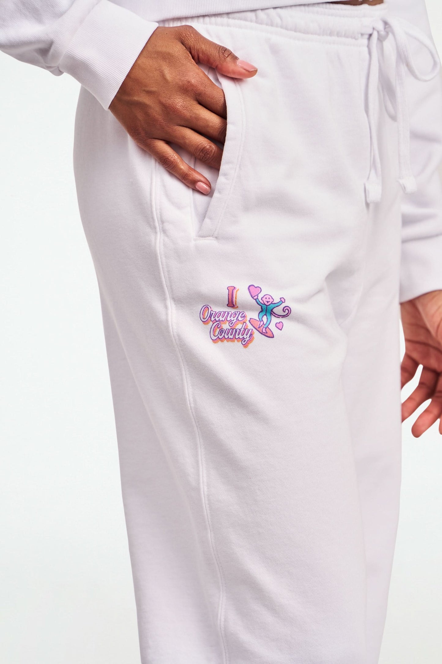 A person stands barefoot in Roller Rabbit O.C. Monkey Sweatpants and a matching hoodie, featuring colorful embroidered and printed graphics near the left pocket. The background is plain white.