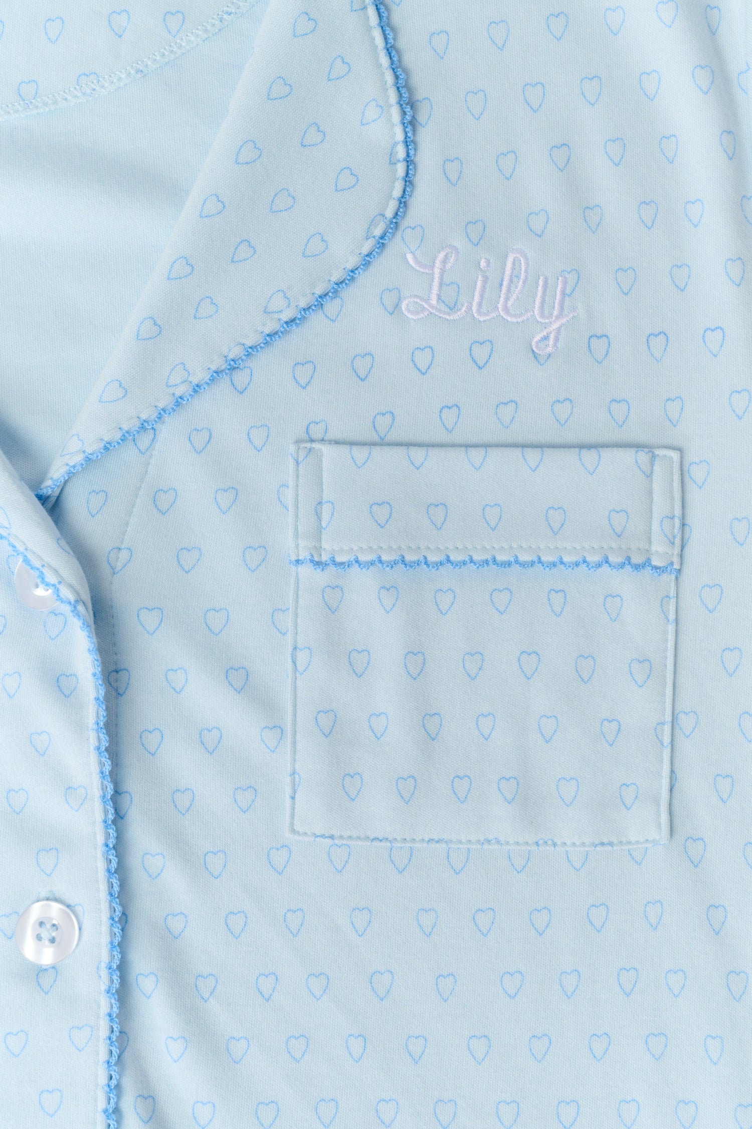 A woman wears Roller Rabbit's Hearts Tonal Wyatt Polo Pajamas—light blue, short-sleeved Pima cotton with a collar and chest pocket—paired with matching ruffled shorts, standing against a plain light background.