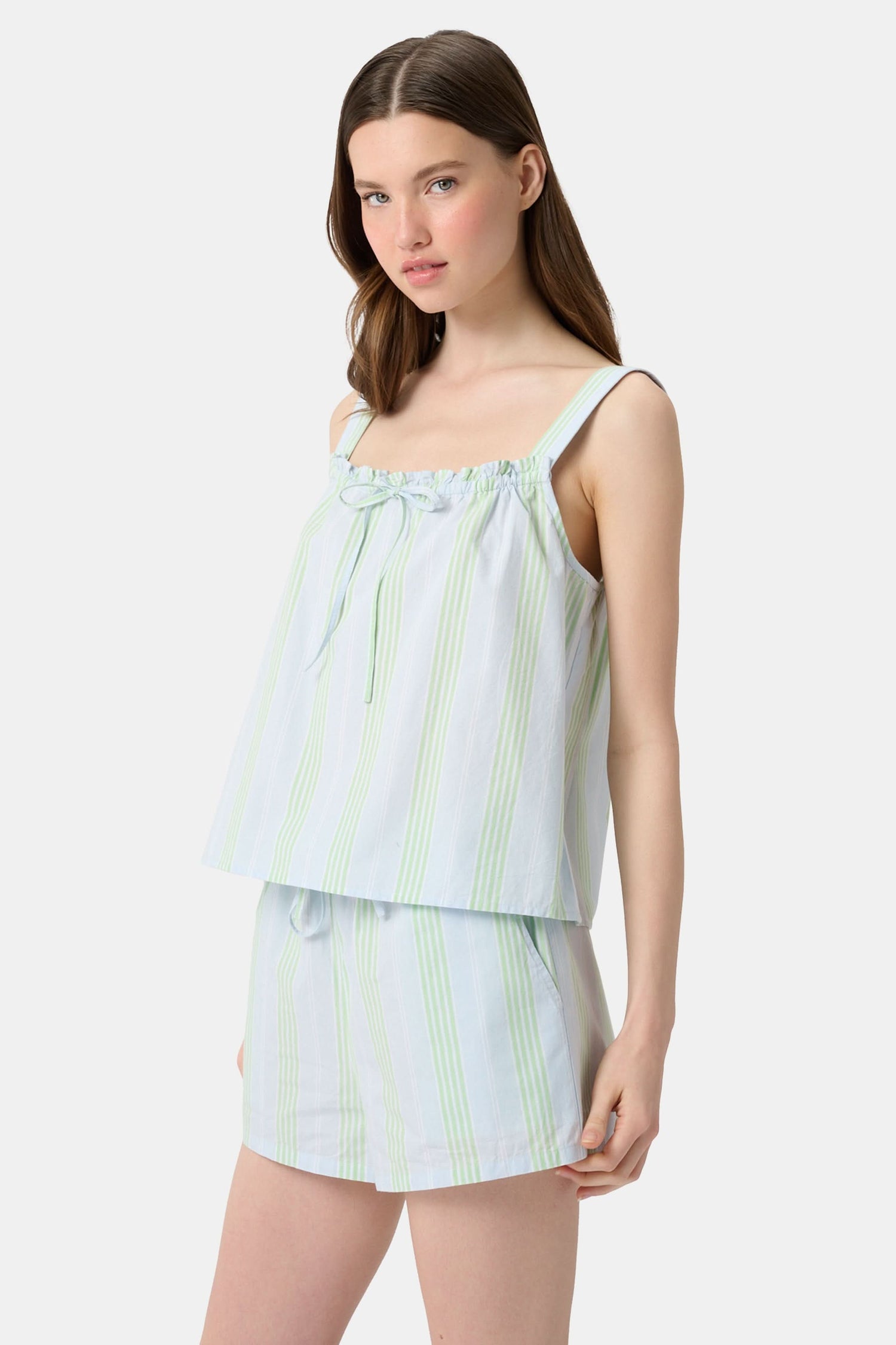 A woman with long brown hair wears the Roller Rabbit Positano Stripe Cooper Set, featuring a loose, sleeveless cotton poplin top with ruffle trim and drawstring, and elastic-waist shorts in white fabric with light green vertical stripes.