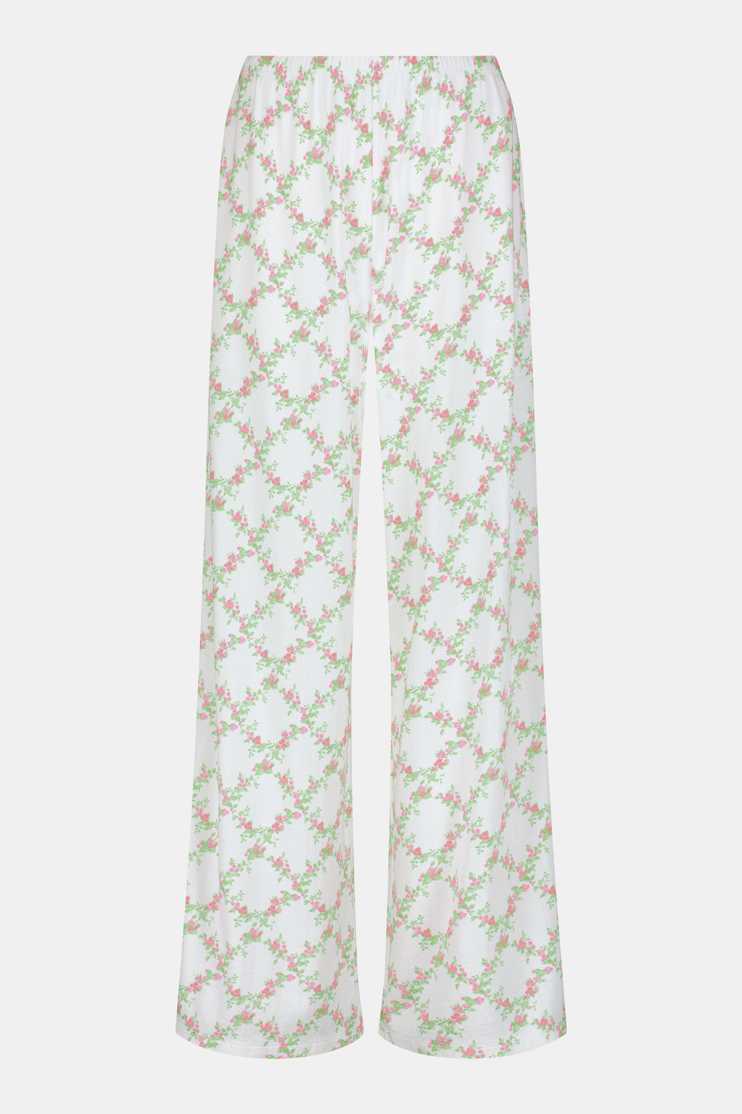 Someone stands against a plain white background wearing Roller Rabbit's Raspberry Garden Monroe Pant—loose white loungewear pants with a pink and green floral print—paired with maroon slippers adorned with white floral details.