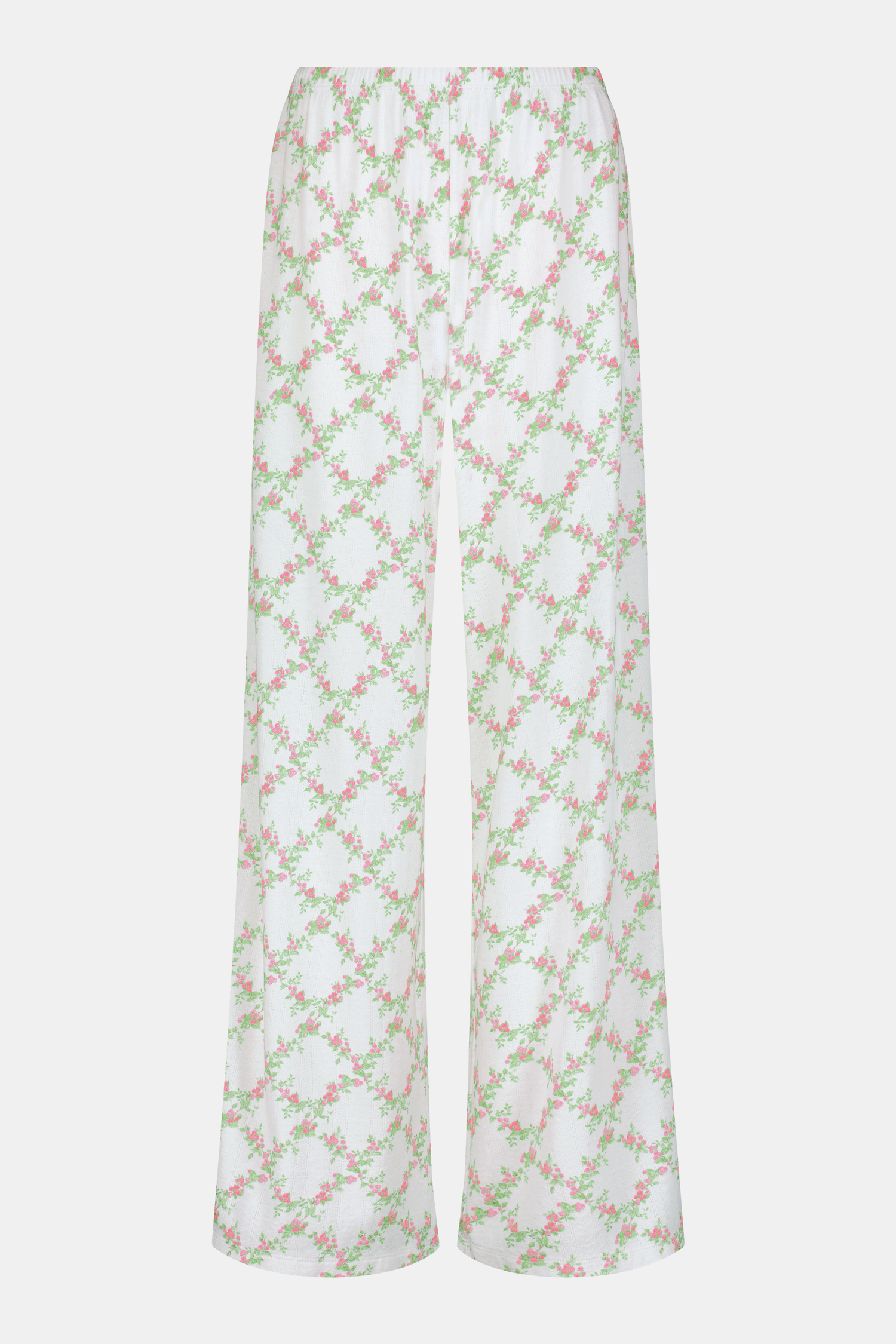 Someone stands against a plain white background wearing Roller Rabbit's Raspberry Garden Monroe Pant—loose white loungewear pants with a pink and green floral print—paired with maroon slippers adorned with white floral details.