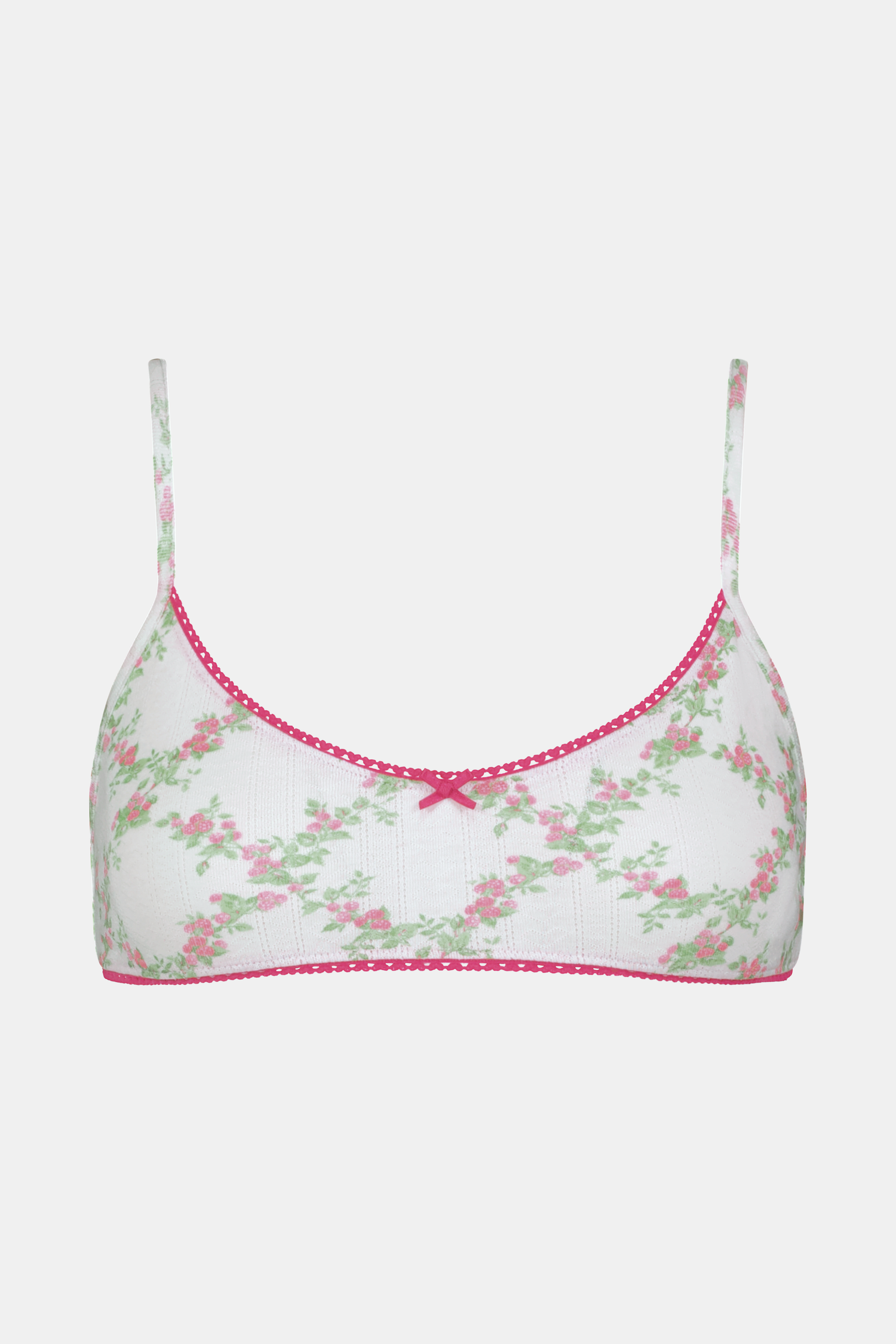 A woman wears the Roller Rabbit Raspberry Garden Ana Scoop Bralette, featuring pink and green floral patterns, pink trim, and a small pink bow. She looks down with one hand behind her head.