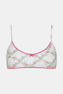 A woman wears the Roller Rabbit Raspberry Garden Ana Scoop Bralette, featuring pink and green floral patterns, pink trim, and a small pink bow. She looks down with one hand behind her head.