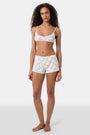 A woman wears the Roller Rabbit Raspberry Garden Ana Scoop Bralette, featuring pink and green floral patterns, pink trim, and a small pink bow. She looks down with one hand behind her head.