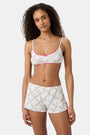 A woman wears the Roller Rabbit Raspberry Garden Ana Scoop Bralette, featuring pink and green floral patterns, pink trim, and a small pink bow. She looks down with one hand behind her head.