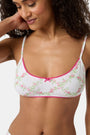 A woman wears the Roller Rabbit Raspberry Garden Ana Scoop Bralette, featuring pink and green floral patterns, pink trim, and a small pink bow. She looks down with one hand behind her head.