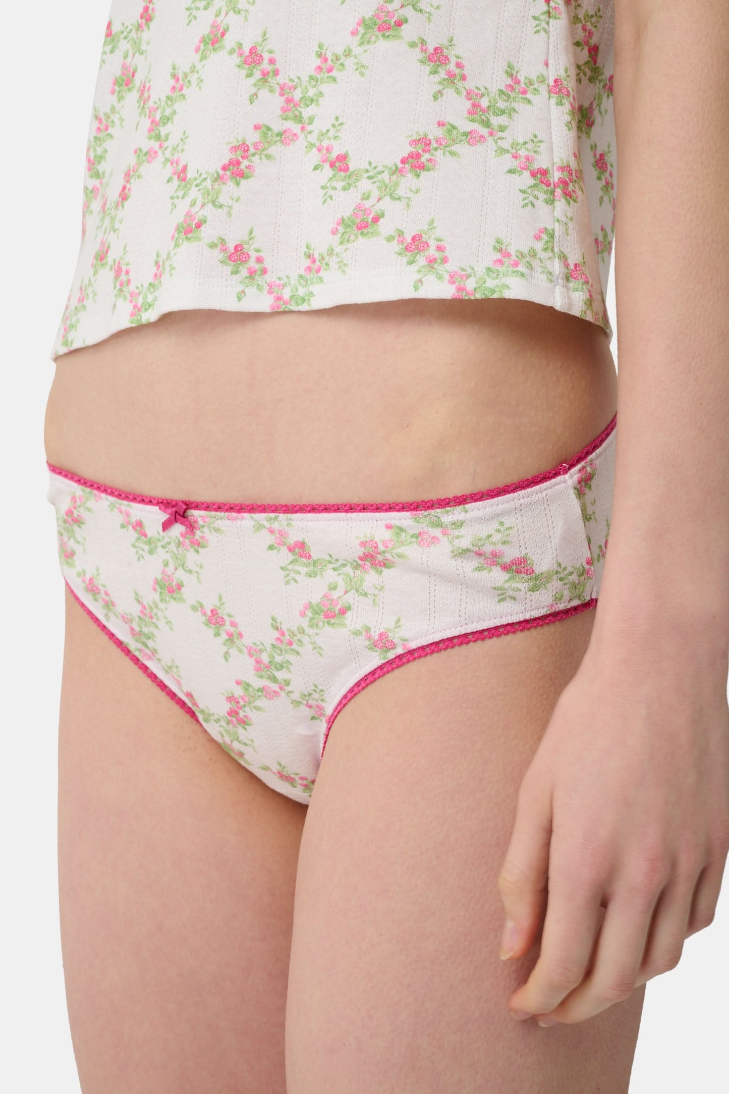 A person wearing the Roller Rabbit Raspberry Garden Gwen Bikini, a matching white camisole and vintage-inspired underwear set with a pink floral pattern and pink trim, shown from mid-torso to upper thighs.