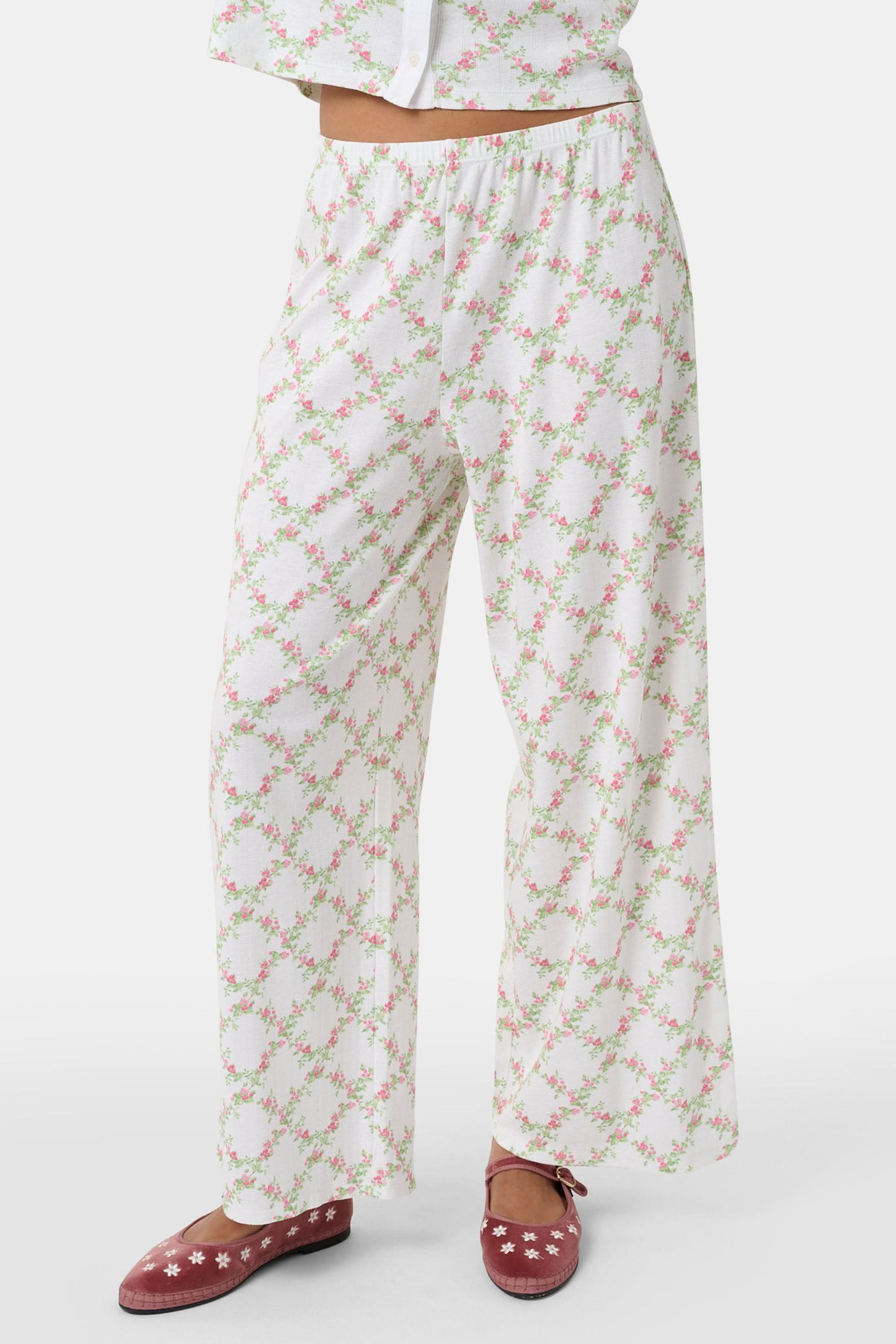 Someone stands against a plain white background wearing Roller Rabbit's Raspberry Garden Monroe Pant—loose white loungewear pants with a pink and green floral print—paired with maroon slippers adorned with white floral details.