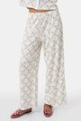 Someone stands against a plain white background wearing Roller Rabbit's Raspberry Garden Monroe Pant—loose white loungewear pants with a pink and green floral print—paired with maroon slippers adorned with white floral details.