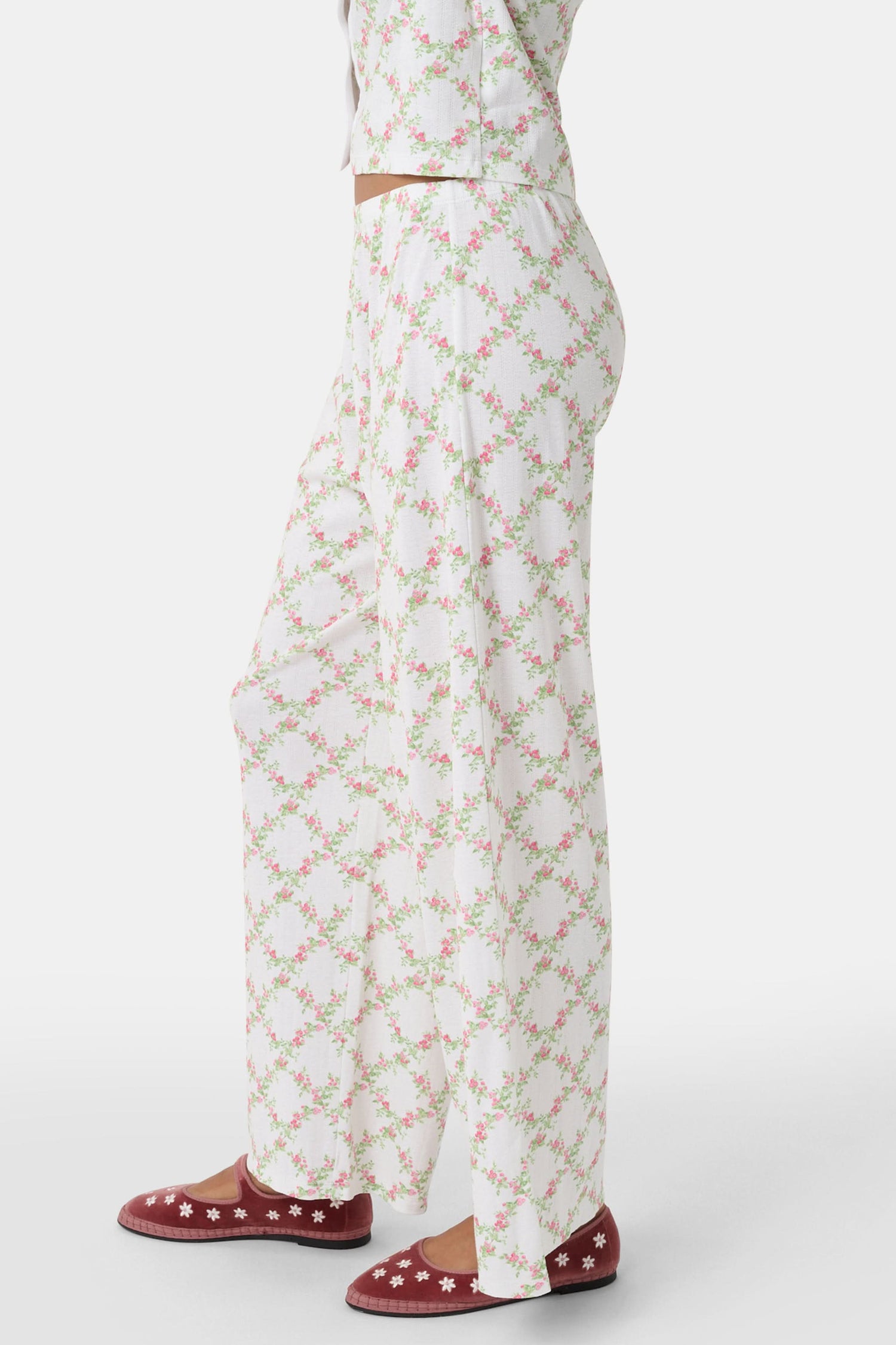 Someone stands against a plain white background wearing Roller Rabbit's Raspberry Garden Monroe Pant—loose white loungewear pants with a pink and green floral print—paired with maroon slippers adorned with white floral details.