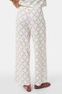 Someone stands against a plain white background wearing Roller Rabbit's Raspberry Garden Monroe Pant—loose white loungewear pants with a pink and green floral print—paired with maroon slippers adorned with white floral details.