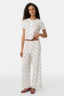 Someone stands against a plain white background wearing Roller Rabbit's Raspberry Garden Monroe Pant—loose white loungewear pants with a pink and green floral print—paired with maroon slippers adorned with white floral details.