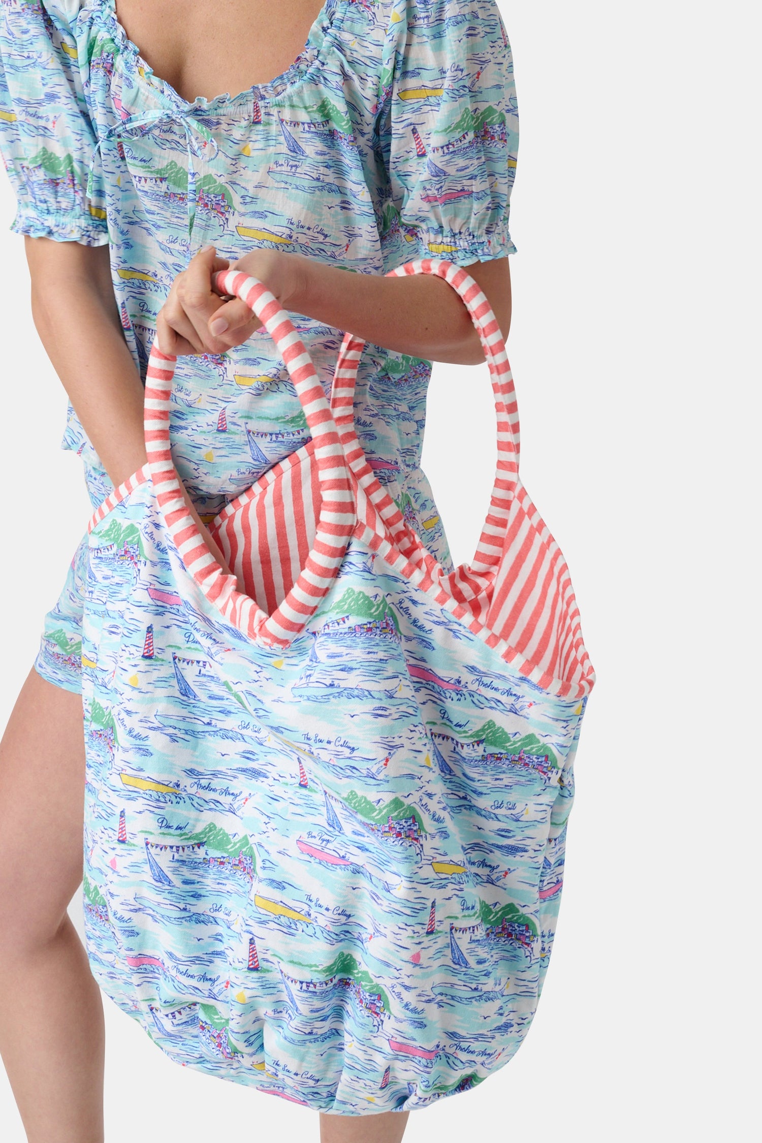 The Regatta Bondi Bag by Roller Rabbit is a large cotton canvas tote with pink and white striped handles, showcasing a vibrant nautical print of boats, lighthouses, and coastal scenery on a white background.