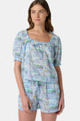 A woman wears the Roller Rabbit Regatta Preston Set, a cotton voile two-piece with a pastel coastal print, featuring a short-sleeve top with gathered neckline and matching shorts, standing against a plain white background.