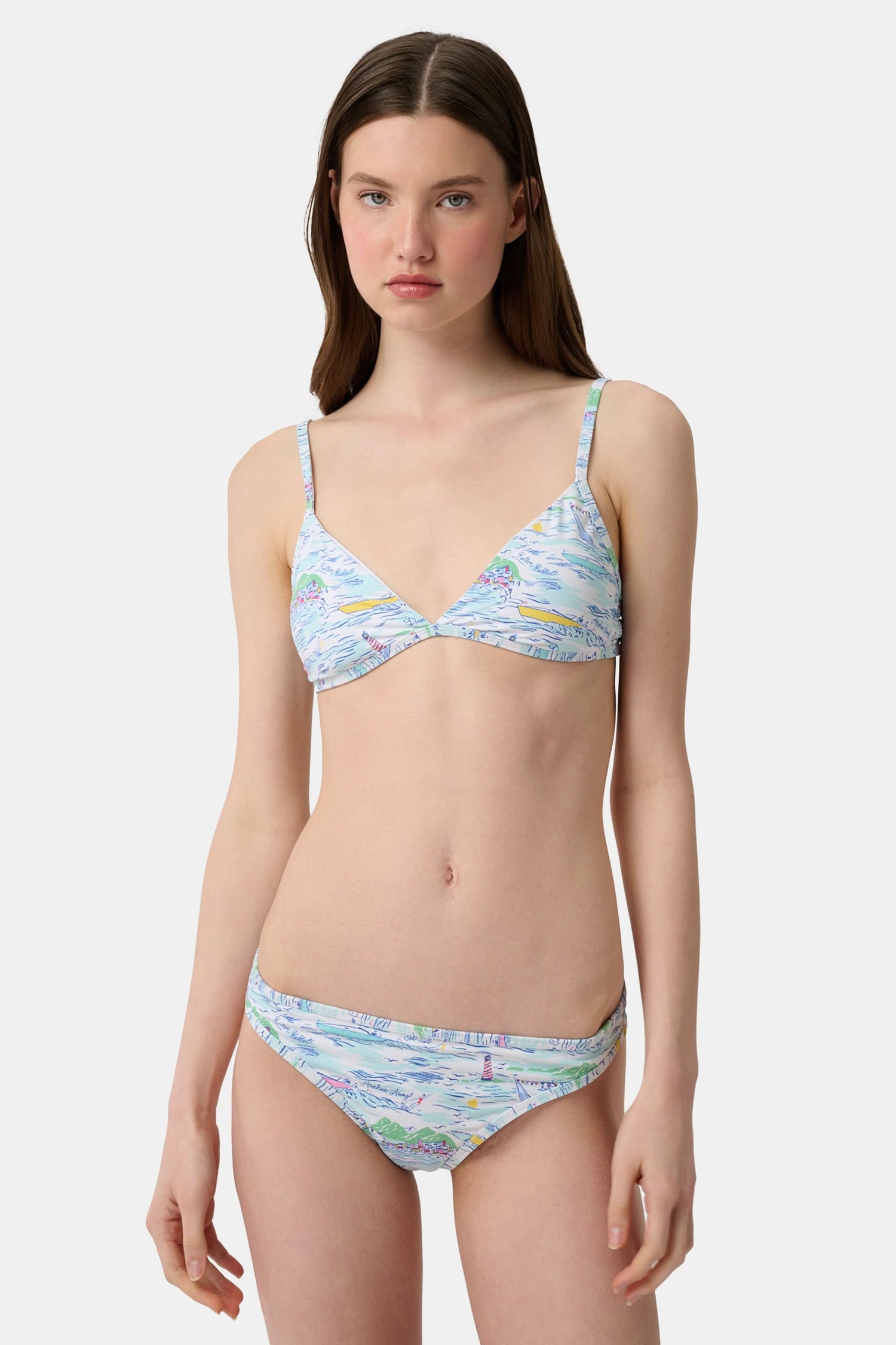 A woman with long brown hair smiles softly, wearing the Regatta Marais Bikini Top by Roller Rabbit in a nautical print, standing against a plain white background.