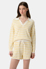 A young woman with light skin and long brown hair wears the Roller Rabbit Striped Rocio Sweater, a pastel yellow, lightweight cotton knit with a wide V-neck and collar. She pairs it with matching drawstring pants and gazes slightly to the side.