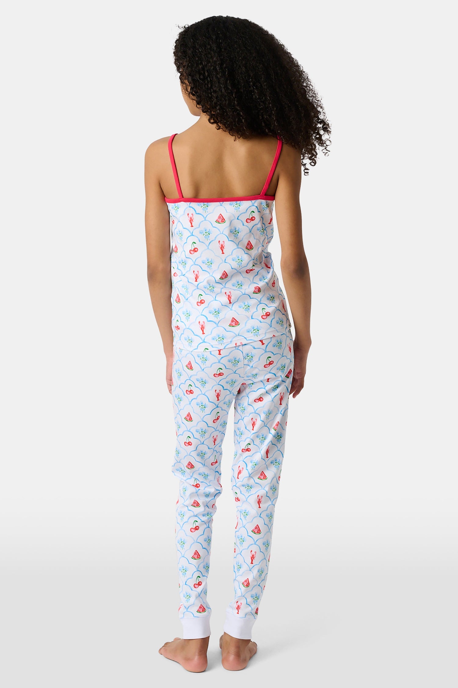 A young girl stands barefoot, smiling against a light background. She wears the Roller Rabbit Kids 4th of Jemina Noa Set—matching white Pima Cotton pajamas with red trim and a colorful flowers, butterflies, and birds print.