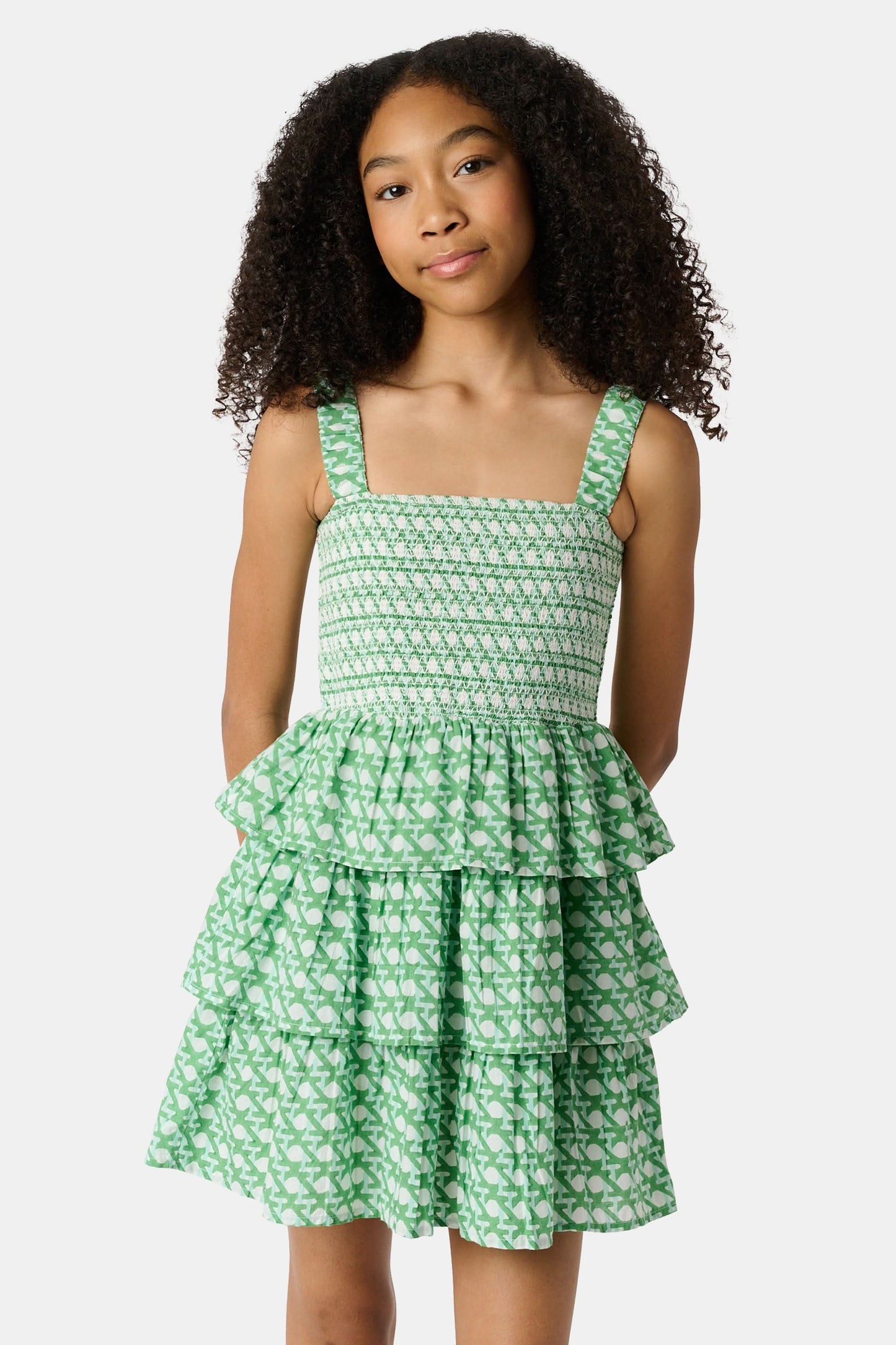 A young girl with curly hair wears the Roller Rabbit Kids Cane Kenia Dress, a green and white patterned cotton tiered dress with wide straps, standing against a plain light background.