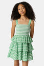 A young girl with curly hair wears the Roller Rabbit Kids Cane Kenia Dress, a green and white patterned cotton tiered dress with wide straps, standing against a plain light background.
