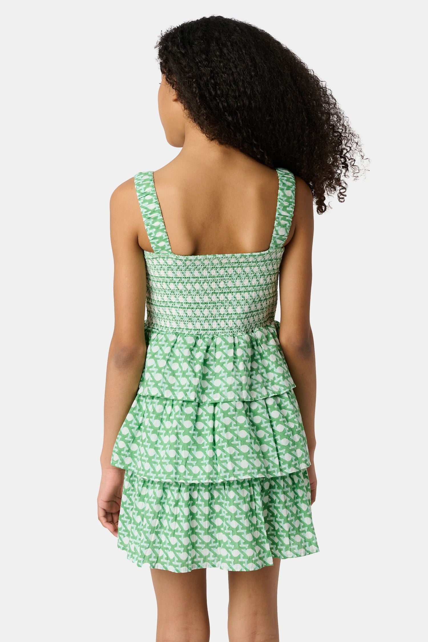A young girl with curly hair wears the Roller Rabbit Kids Cane Kenia Dress, a green and white patterned cotton tiered dress with wide straps, standing against a plain light background.