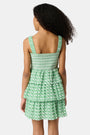 A young girl with curly hair wears the Roller Rabbit Kids Cane Kenia Dress, a green and white patterned cotton tiered dress with wide straps, standing against a plain light background.