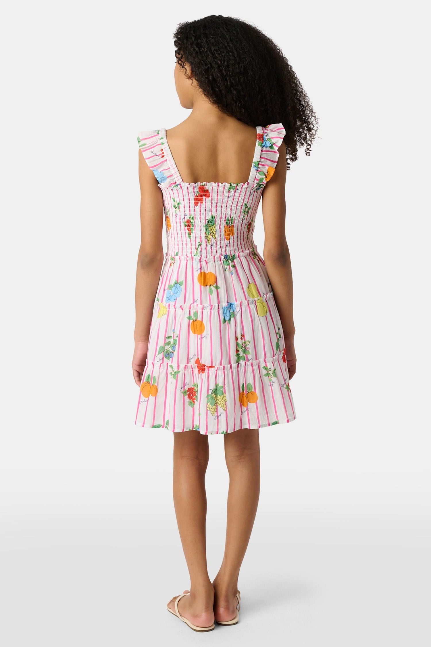 A young girl with straight brown hair wears the Roller Rabbit Kids Fruits D'Ete Ceri Dress, a pink and white smocked sundress with ruffled sleeves, and white sandals, standing and smiling against a light gray background.