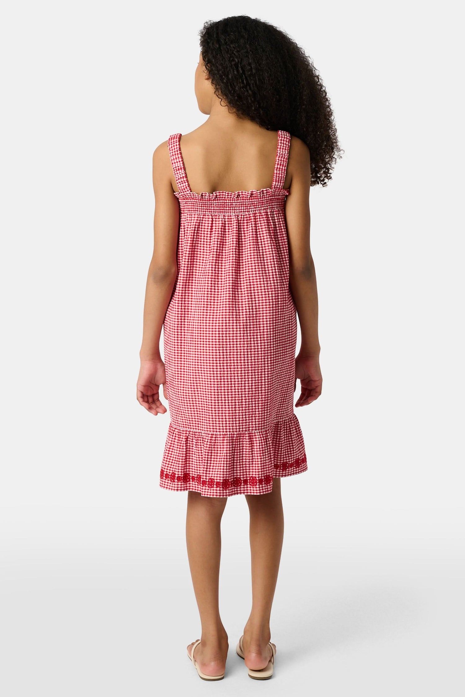 A young girl in the Roller Rabbit Kids Gingham Teagan Dress with an embroidered hem stands barefoot on a boat deck by the water, sunlight shimmering on the surface.