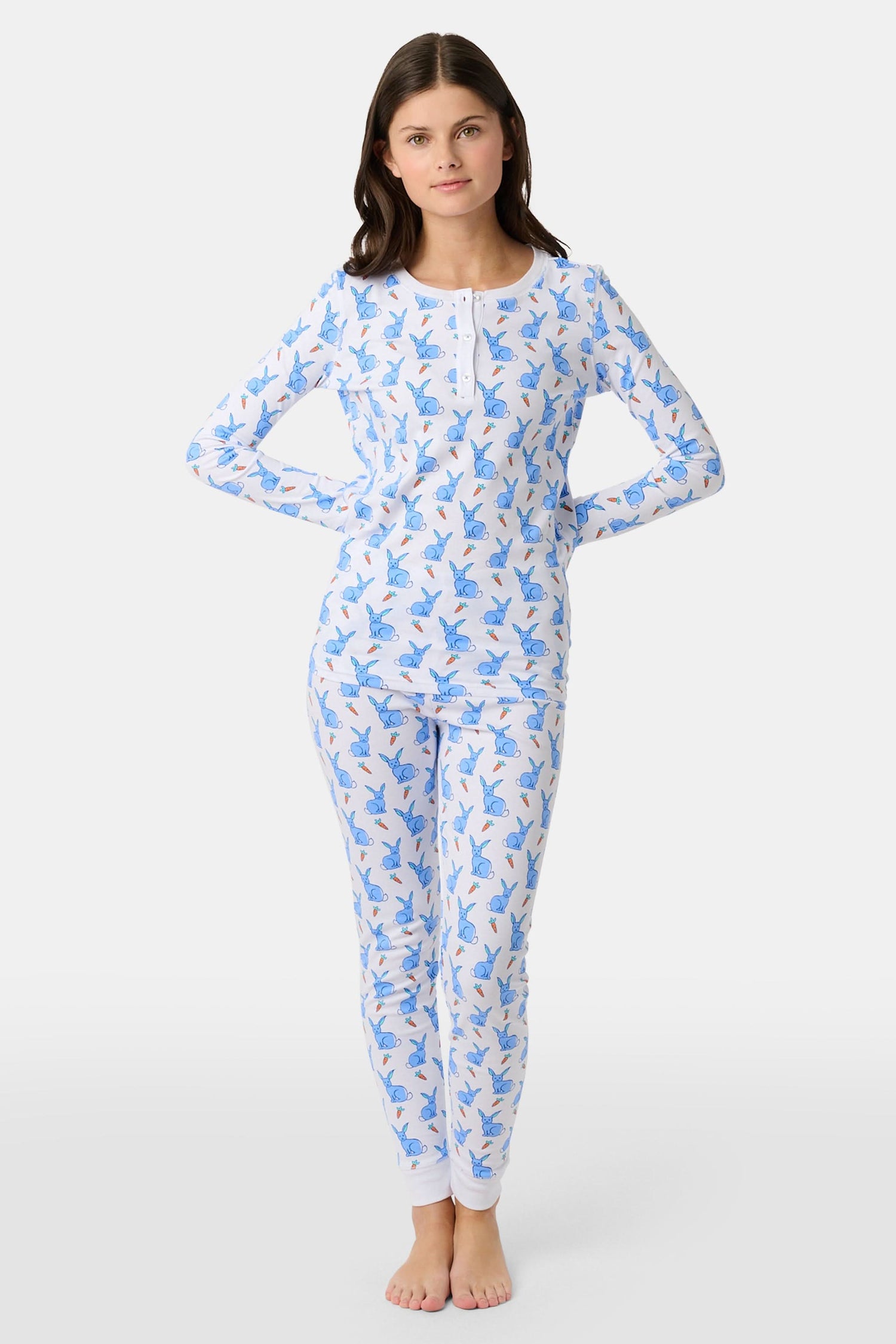 A young child with curly hair stands barefoot in white Kids Romy Rabbit Pajamas by Roller Rabbit, featuring a blue bunny print, looking at the camera and touching their mouth with one finger.