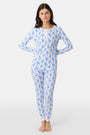 A young child with curly hair stands barefoot in white Kids Romy Rabbit Pajamas by Roller Rabbit, featuring a blue bunny print, looking at the camera and touching their mouth with one finger.