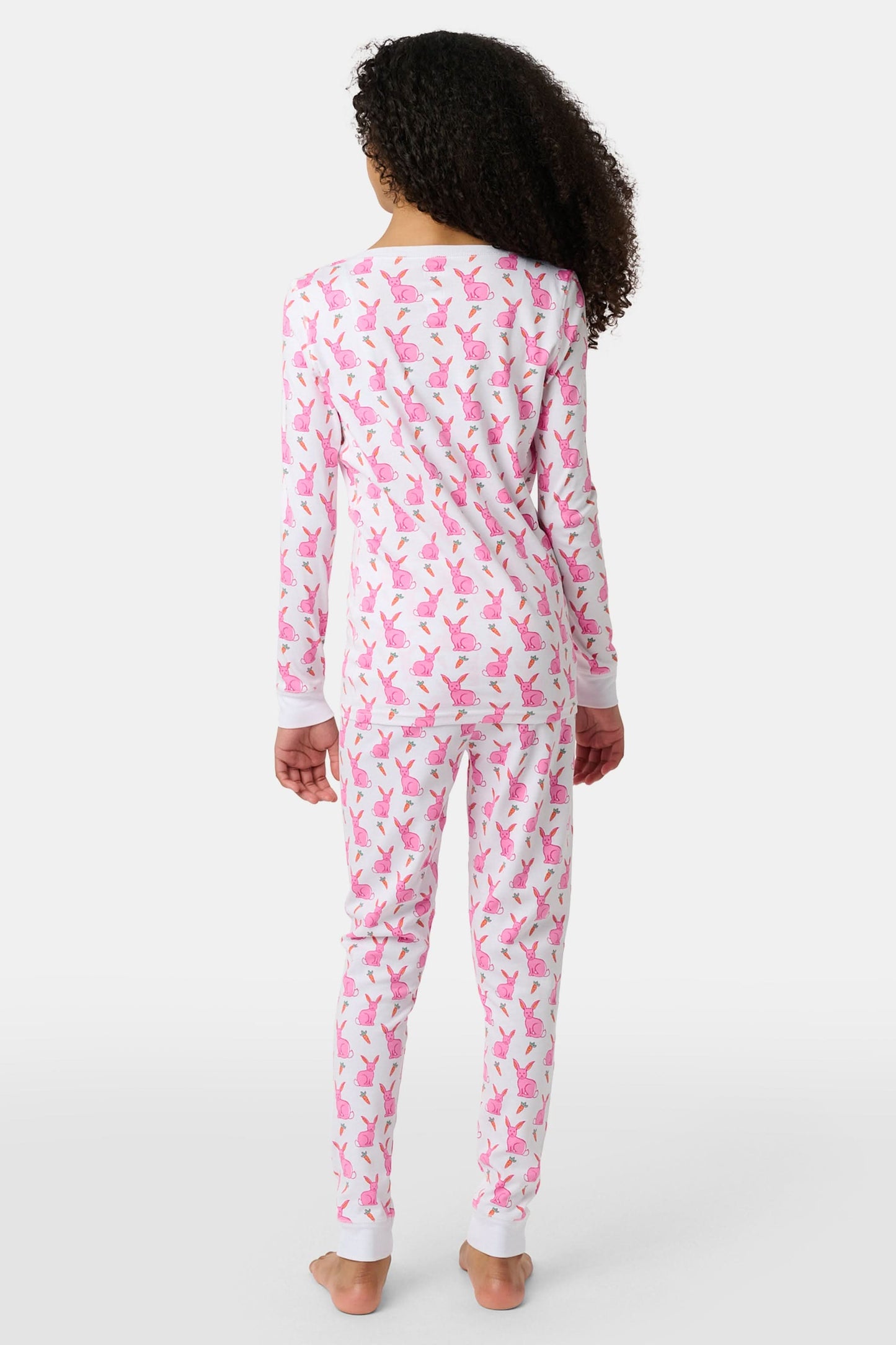 A young girl smiles barefoot in Roller Rabbit’s Kids Romy the Rabbit Pajamas, a soft Pima cotton long-sleeve set with a playful rabbit print; her brown hair frames her face against a simple, light background.